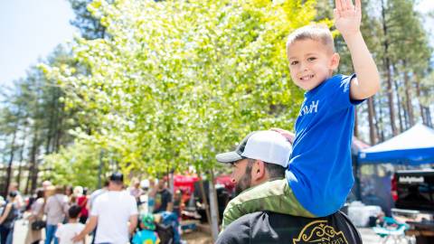 A father carries a child on his shoulders through a busy festival. The little boy turns and waves at the camera